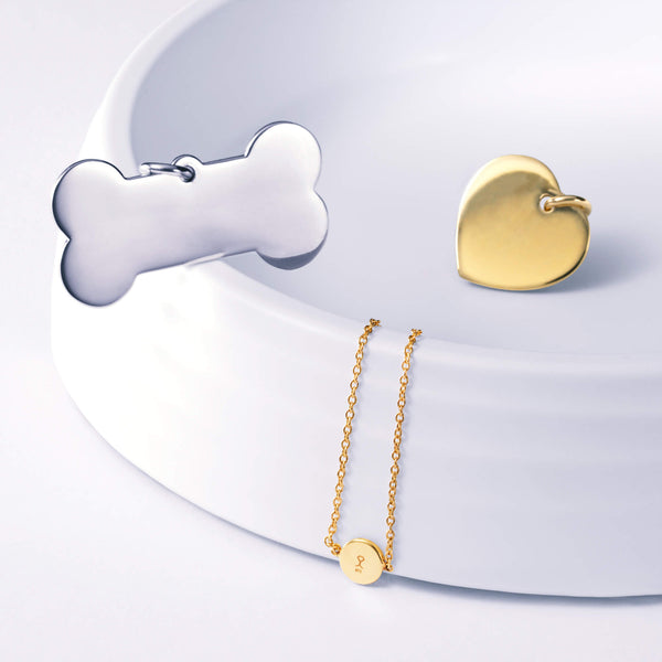 3 jewellery pieces styled together on and around a white dog bowl. A silver bone shape name pendant, a gold heart name pendant and a gold round companion bracelet