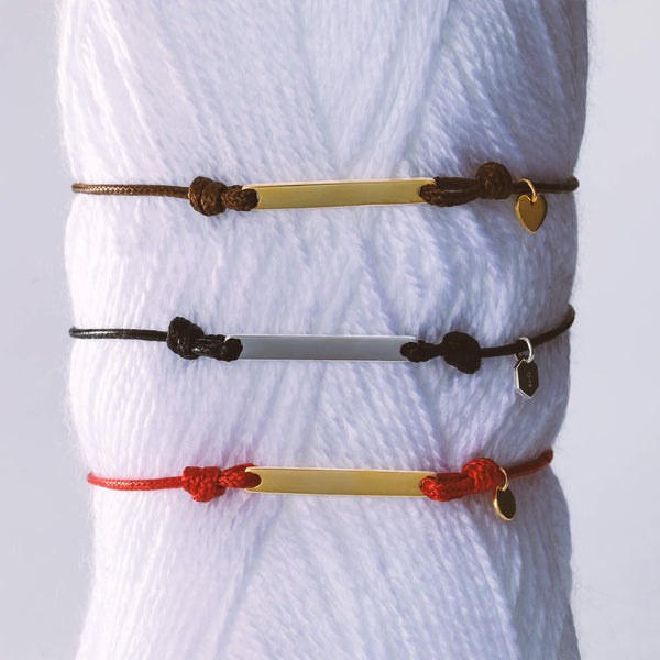 3 companion cord bracelets fastened around a vertical piece of rolled white yarn. Displaying two gold bracelets on a brown and a red cord, and one silver on a black cord