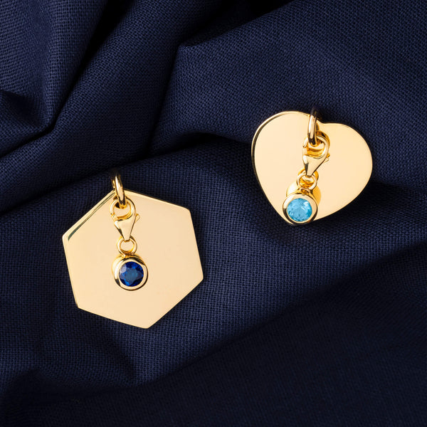 Two styled yellow gold name ID tag pendants with two birthstone pendants attached to their fronts, lying on blue cloth